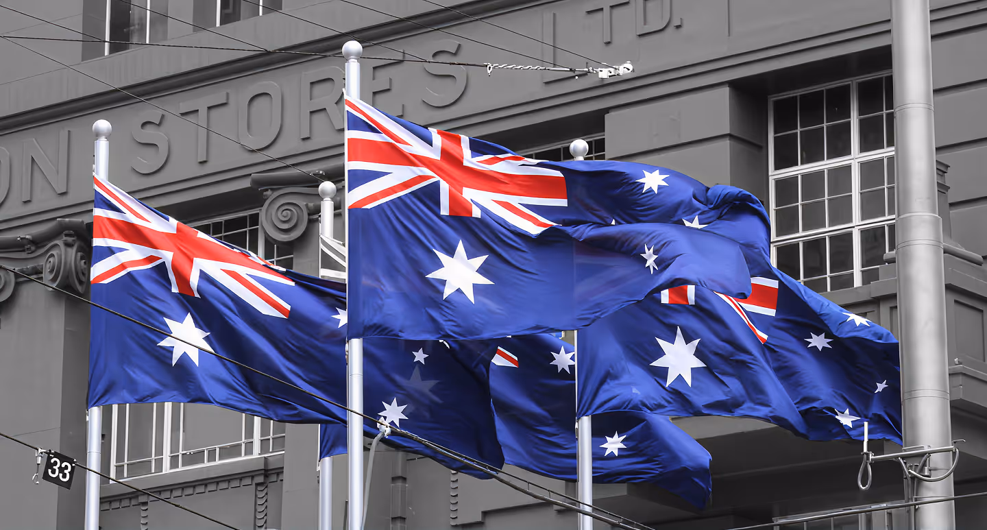 Multiple Australian flags waving on flagpoles in front of a gray building.