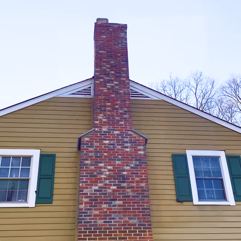 Red and brown brick chimney on a yellow house with green window shutters.