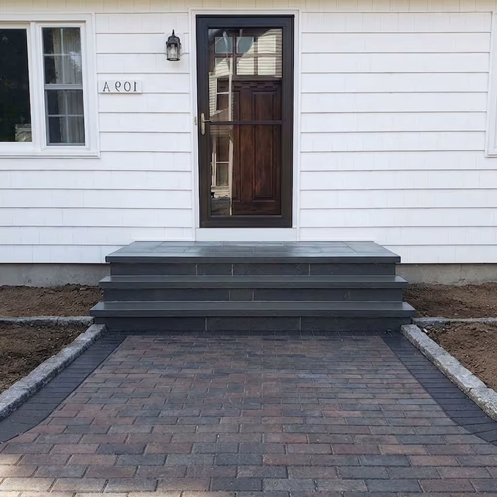 Three wide stone steps leading to a stone porch with cobblestone ground and potted plants on either side.