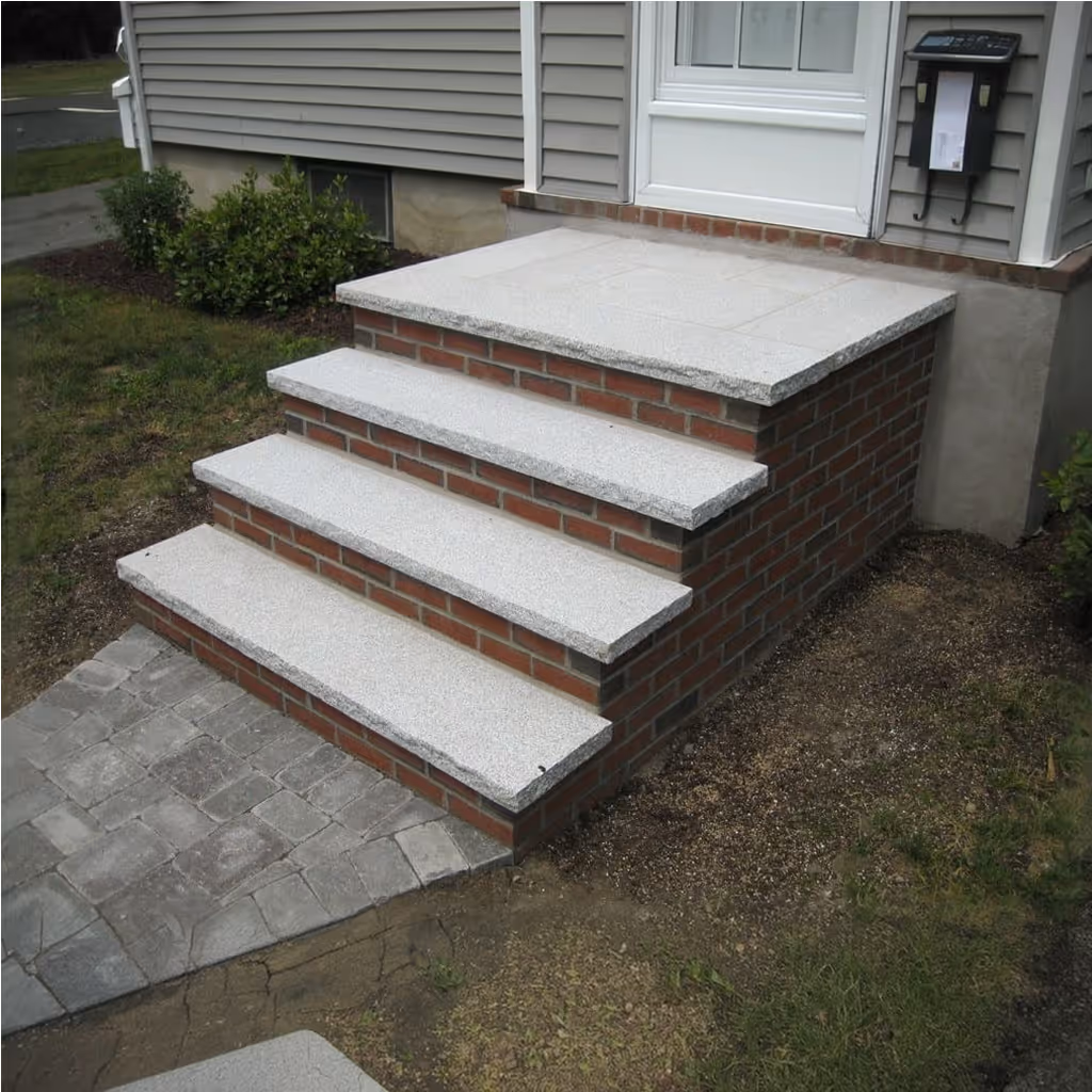 Three wide stone steps leading to a stone porch with cobblestone ground and potted plants on either side.