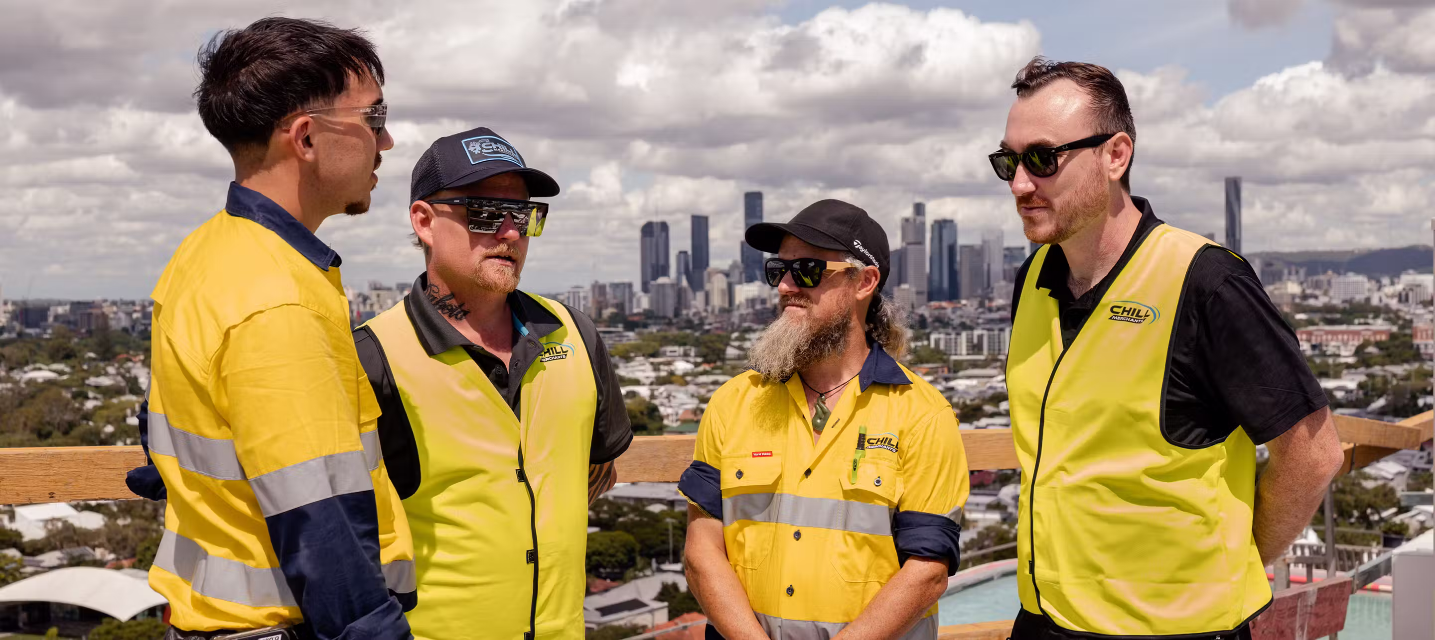 Chill Merchants team members in branded hi-vis and workwear having a discussion with Brisbane city skyline in the background
