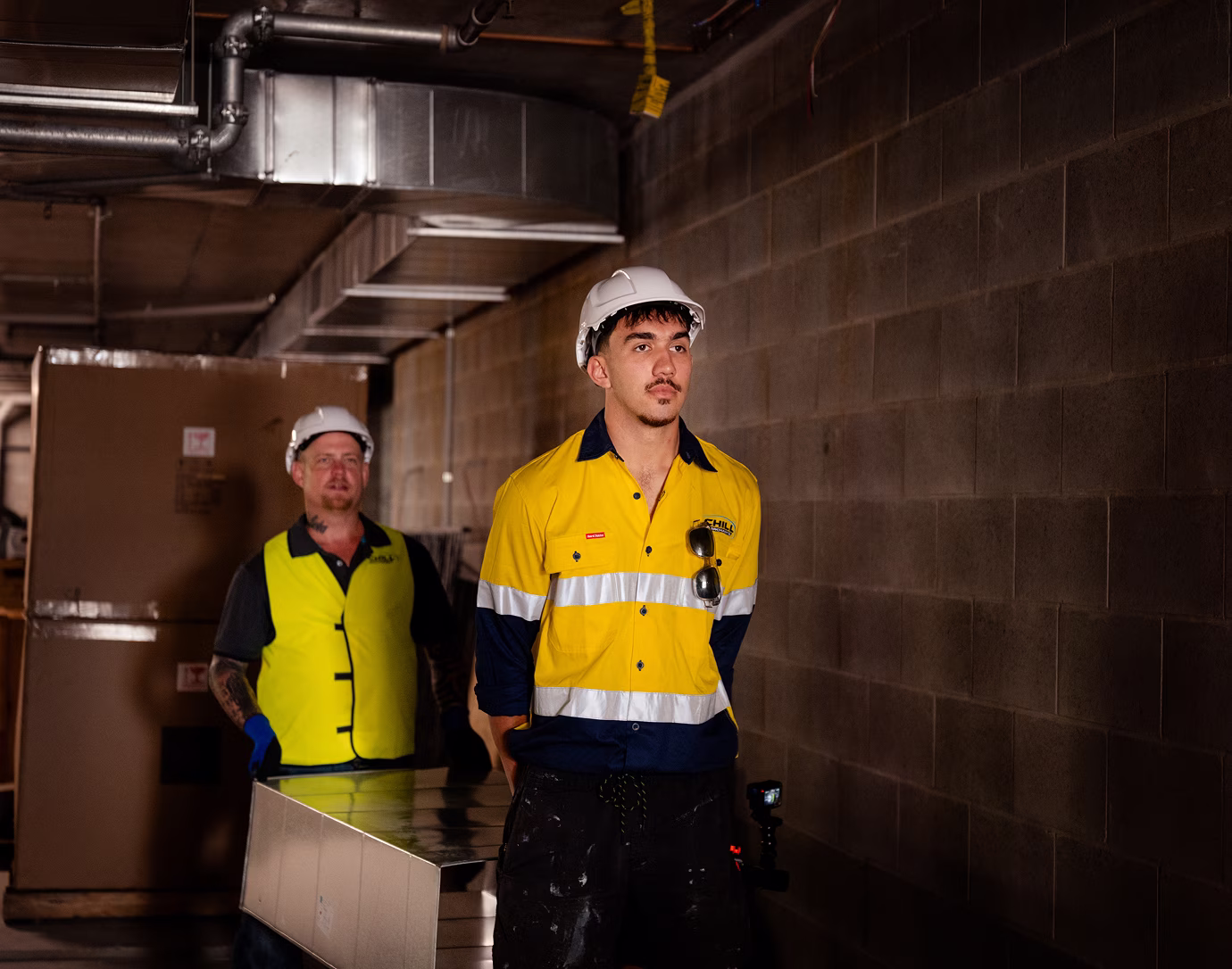 Two Chill Merchants technicians in hi-vis and hard hats walking through a commercial building basement plant room corridor