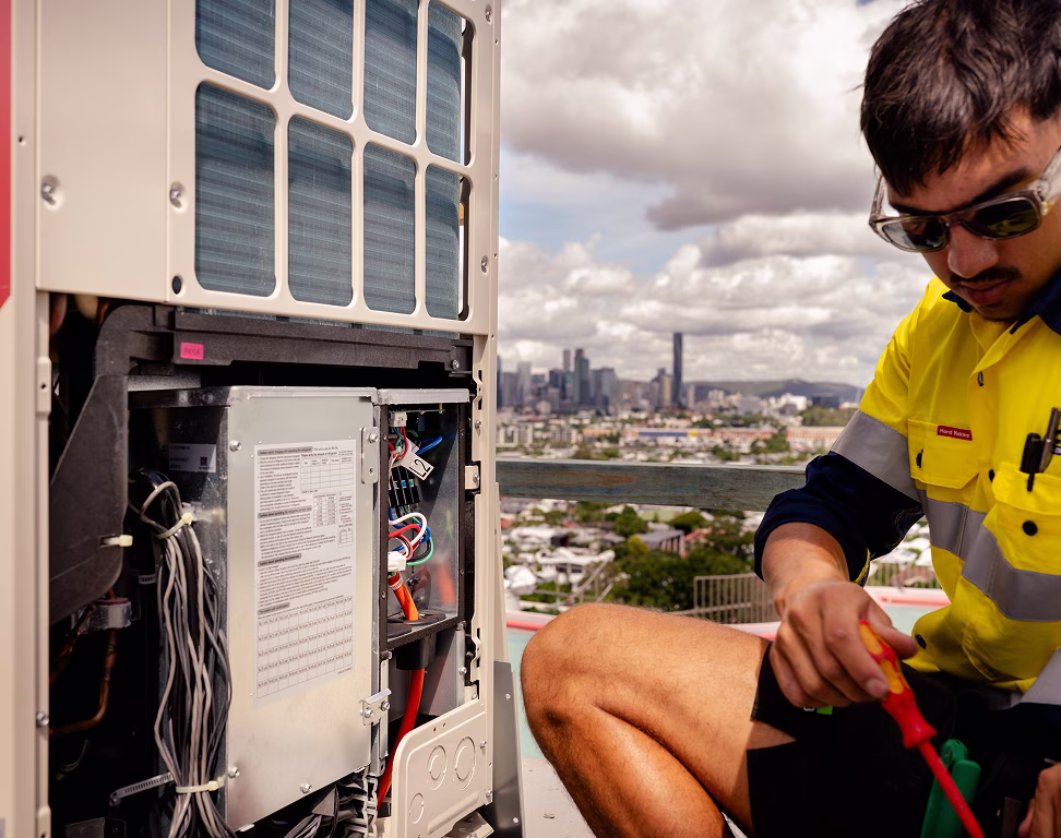 Two Chill Merchants technicians in hi-vis and sunglasses servicing the internal wiring of a Mitsubishi Electric condenser unit on a rooftop with Brisbane skyline behind