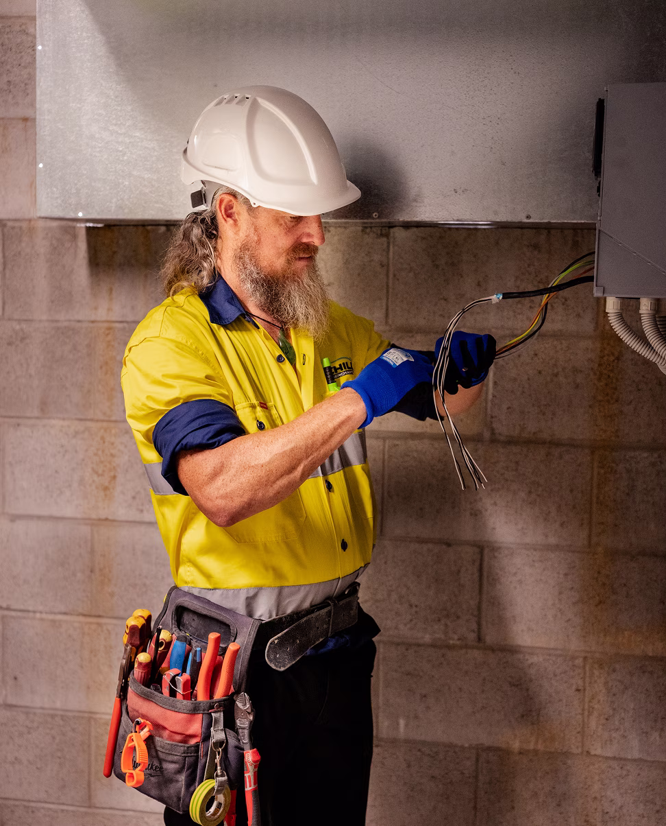 Senior Chill Merchants technician in hi-vis, hard hat, and tool belt pulling electrical cables through a conduit in a commercial plant room