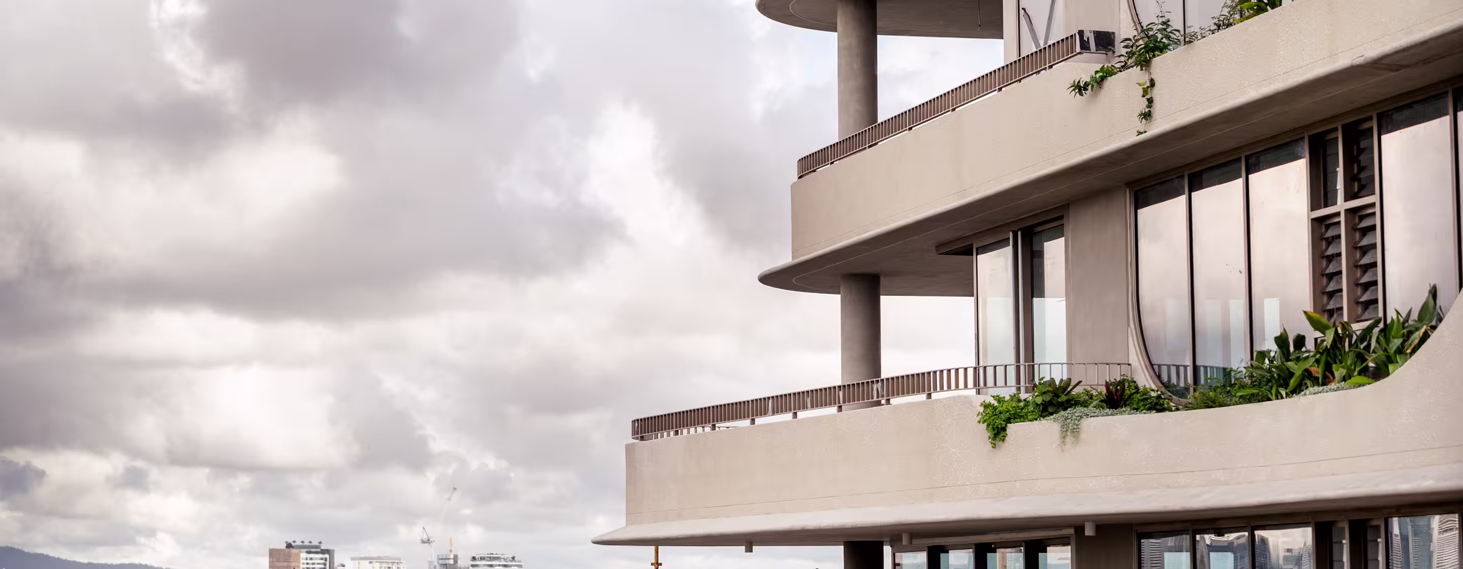 Modern building balcony with curved edges, glass windows, green plants, and cloudy sky background.