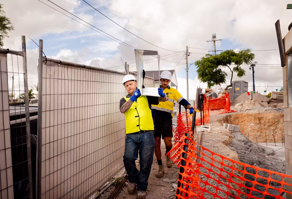 Two construction workers wearing white helmets and yellow vests carrying a metal chair on a construction site with orange safety fencing.