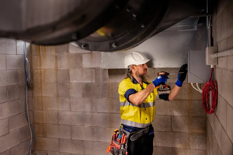 Chill Merchants technician in hi-vis and hard hat using a cordless drill to install ductwork fittings in a tiled commercial space