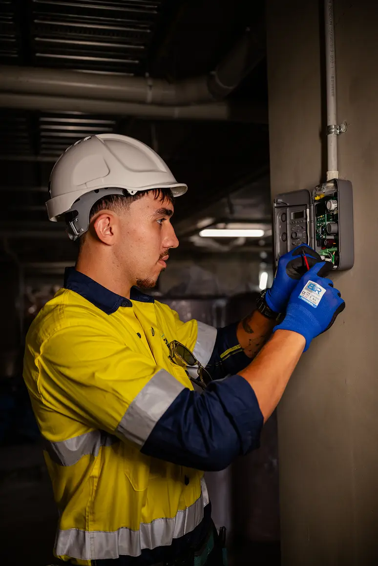 Chill Merchants apprentice in hi-vis, hard hat, and blue gloves using a multimeter to test electrical connections on HVAC equipment in a plant room