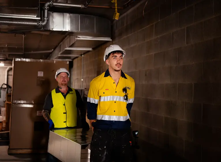 Two male workers wearing yellow safety vests and white helmets, standing in a dimly lit industrial or warehouse space.