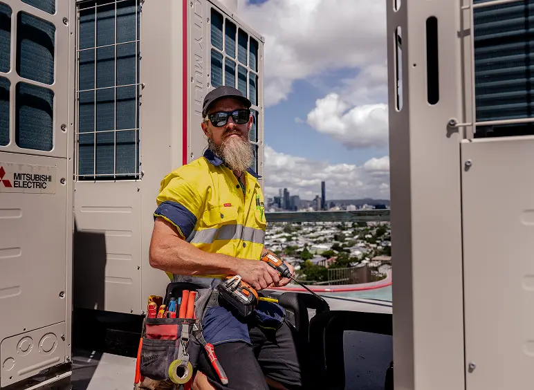 Senior Chill Merchants technician in hi-vis sitting beside a Mitsubishi Electric rooftop condenser unit with Brisbane city skyline in the background
