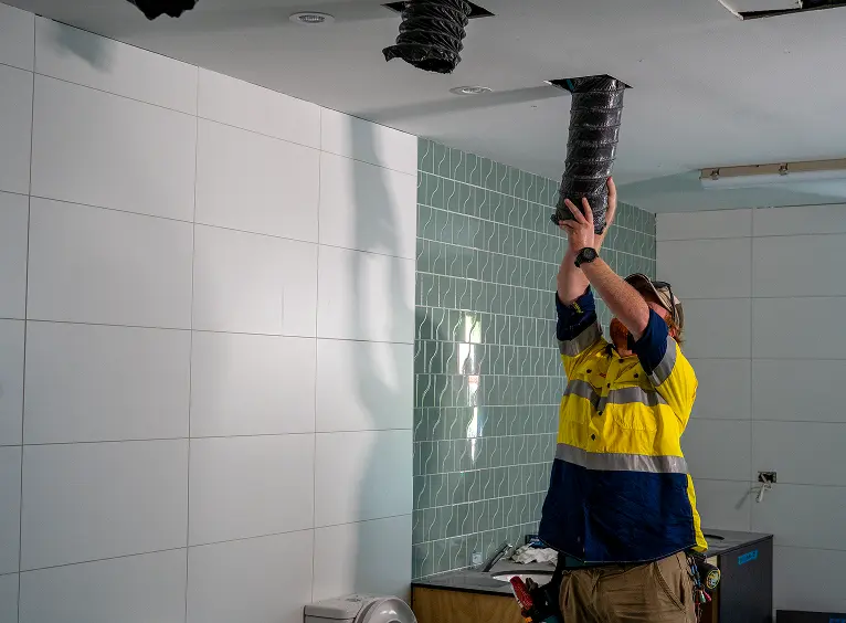 Worker in a high-visibility shirt installing or inspecting black flexible ducting on a ceiling in a tiled room.
