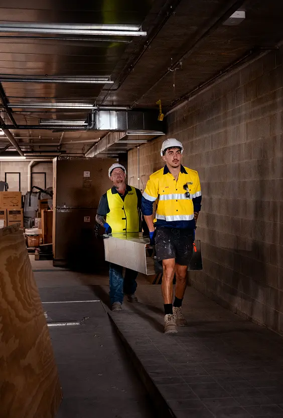 Two Chill Merchants technicians in hi-vis and hard hats carrying a duct section through a commercial building basement corridor