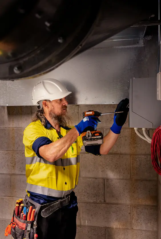 Chill Merchants technician in hi-vis, hard hat, and tool belt using a cordless drill to secure ductwork brackets to a block wall