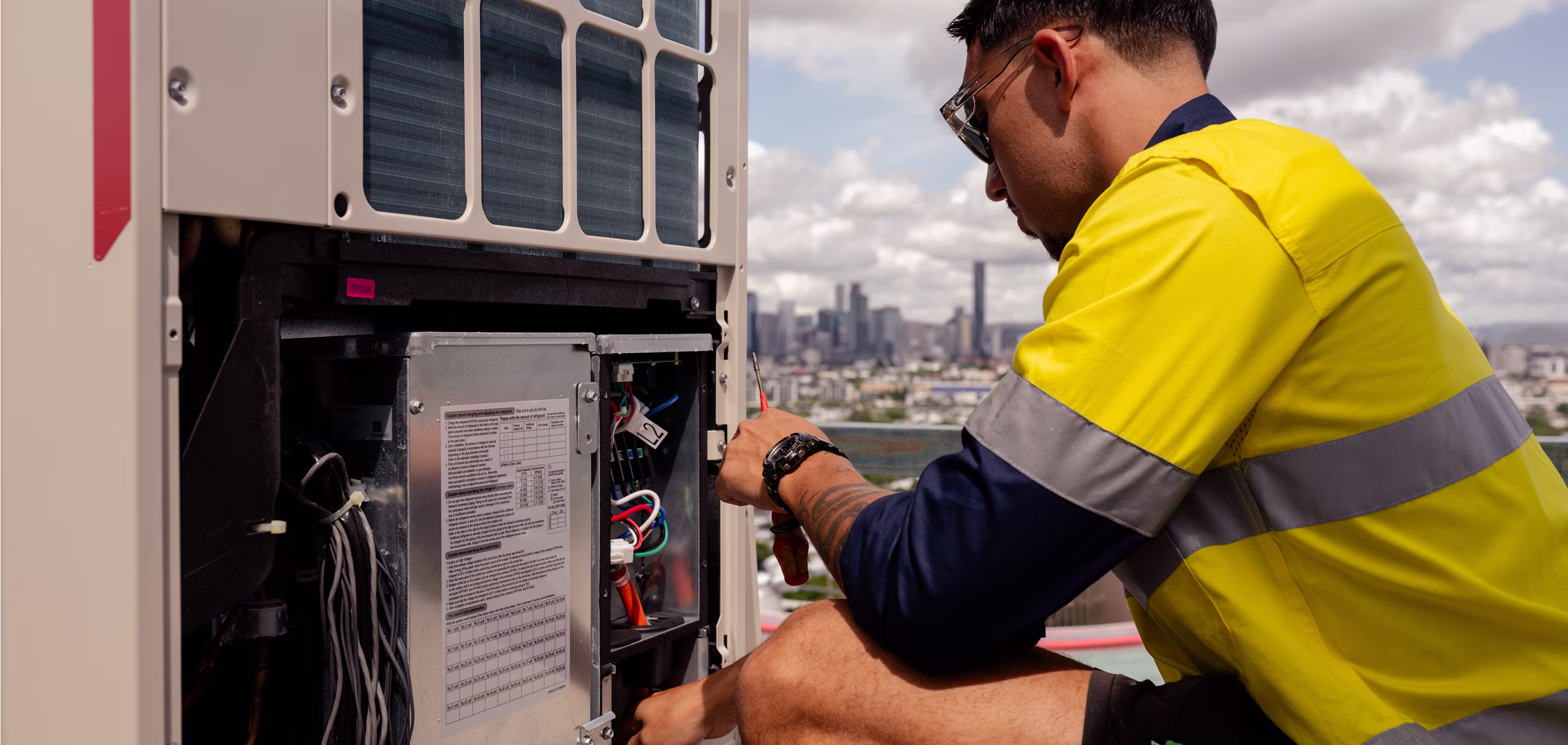 HVAC technician servicing a commercial air conditioning electrical control panel with Brisbane city skyline in the background