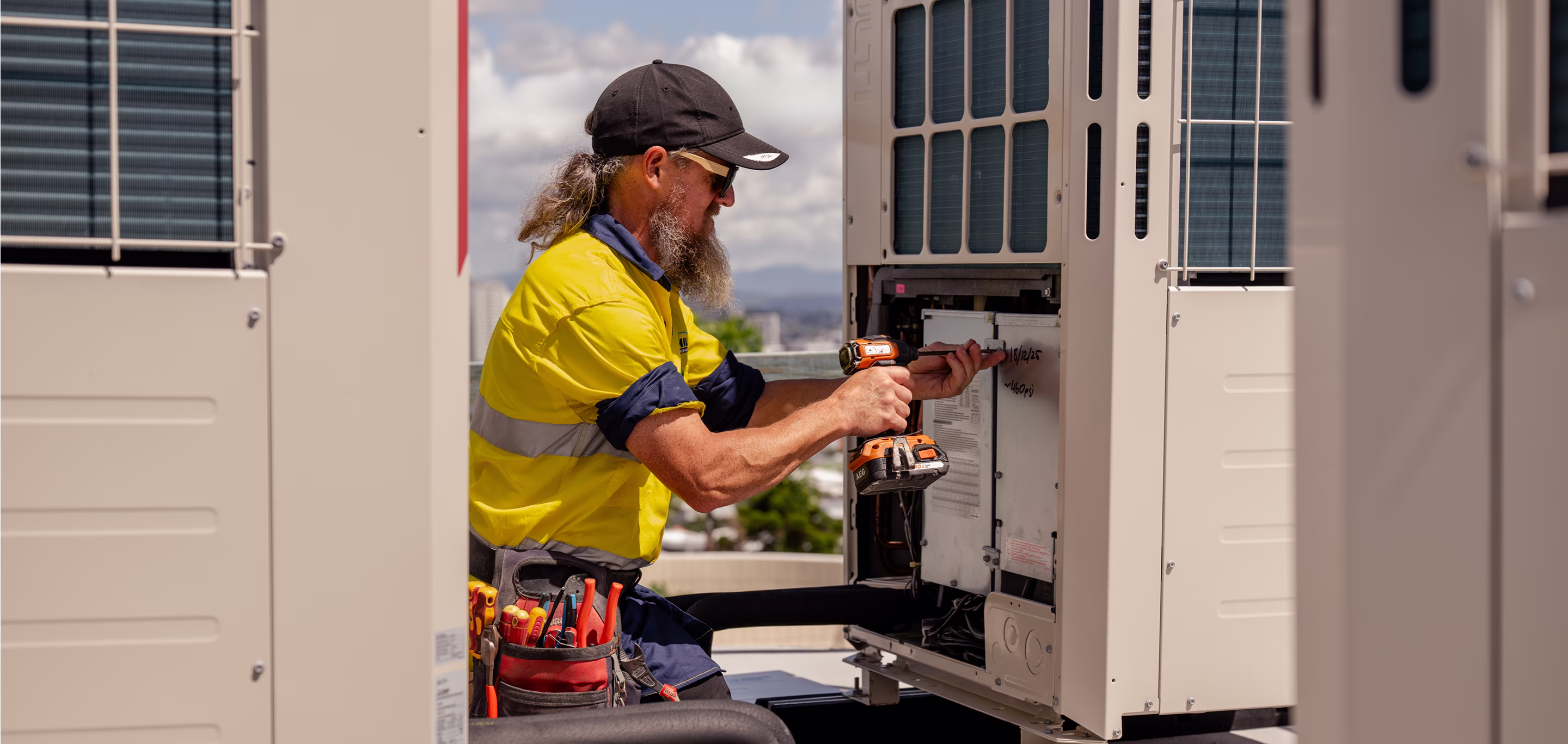HVAC technician in hi-vis workwear using a cordless drill to commission an outdoor electrical panel on a commercial rooftop