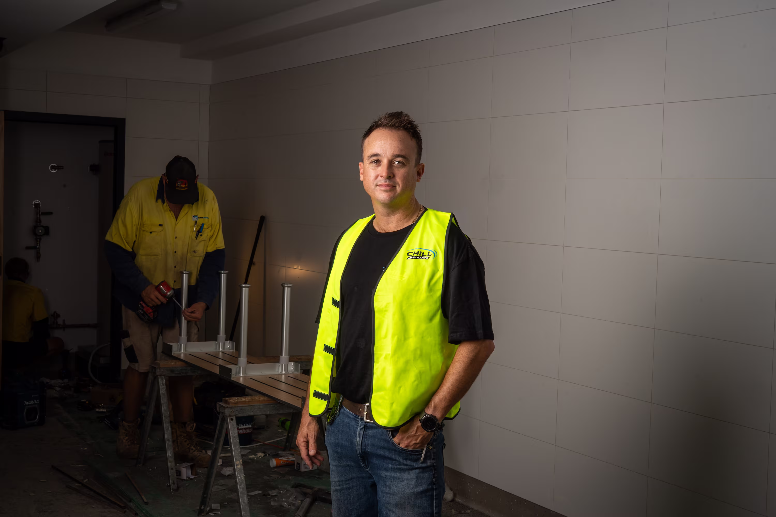 Man in a yellow safety vest stands in a workroom while another worker uses a drill on a table.