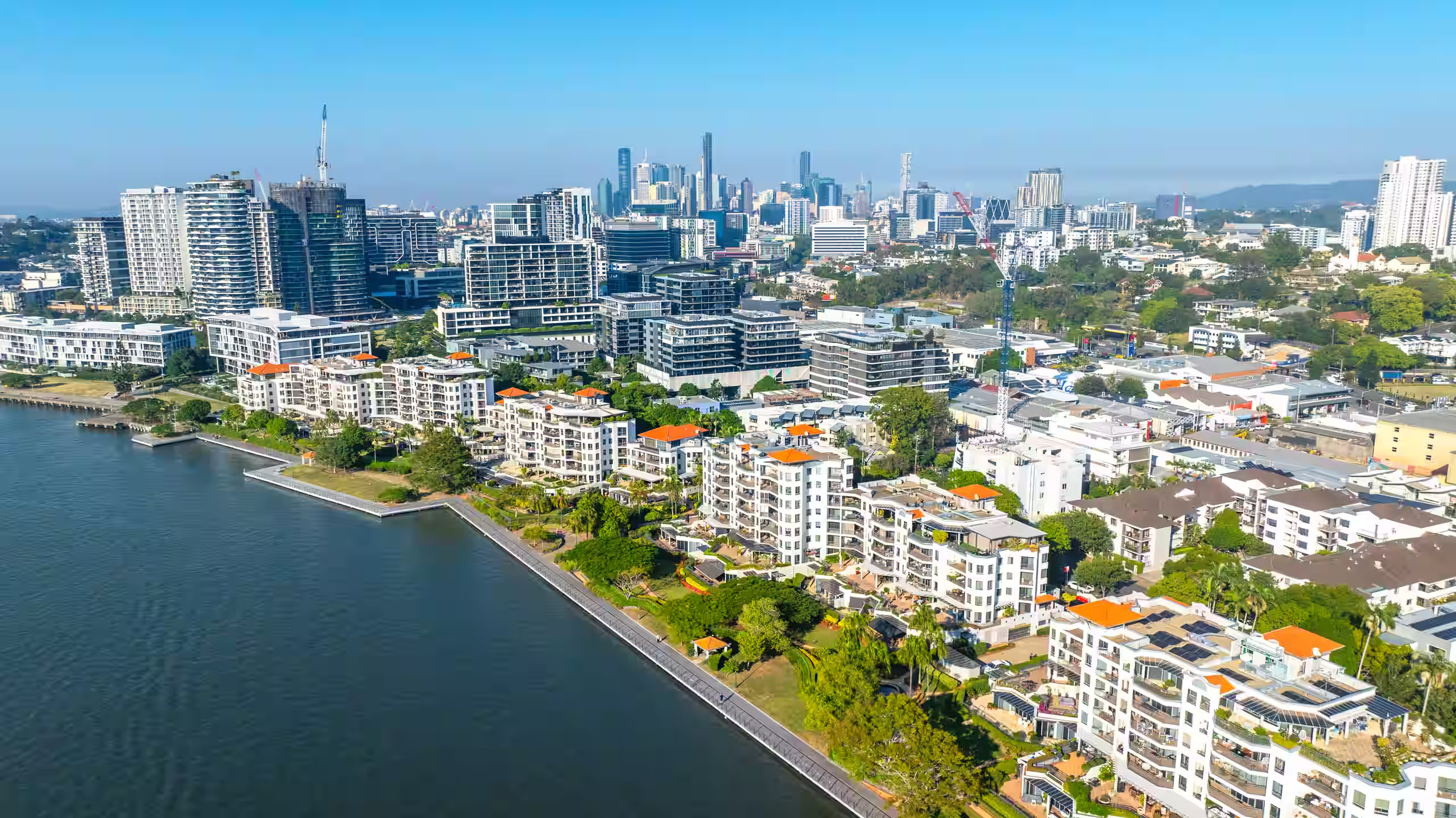 Waterfront multi-residential buildings with orange roofs and city skyline in the background on a clear day.