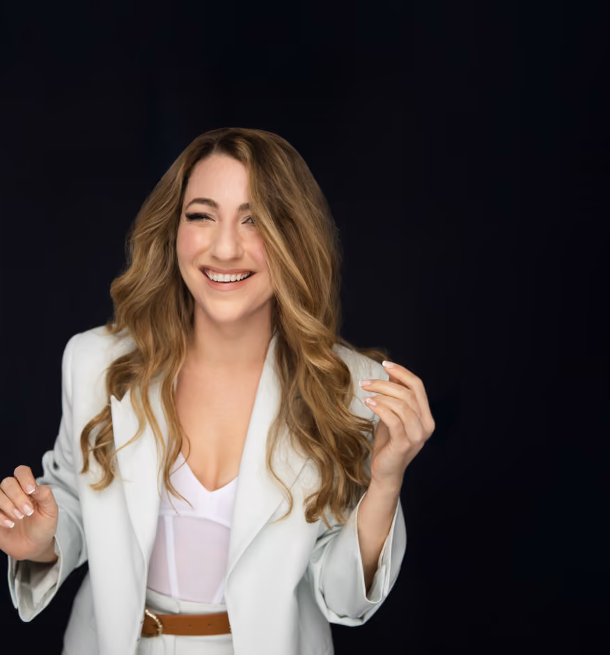 Smiling woman with long wavy hair wearing a white blazer and standing against a black background.