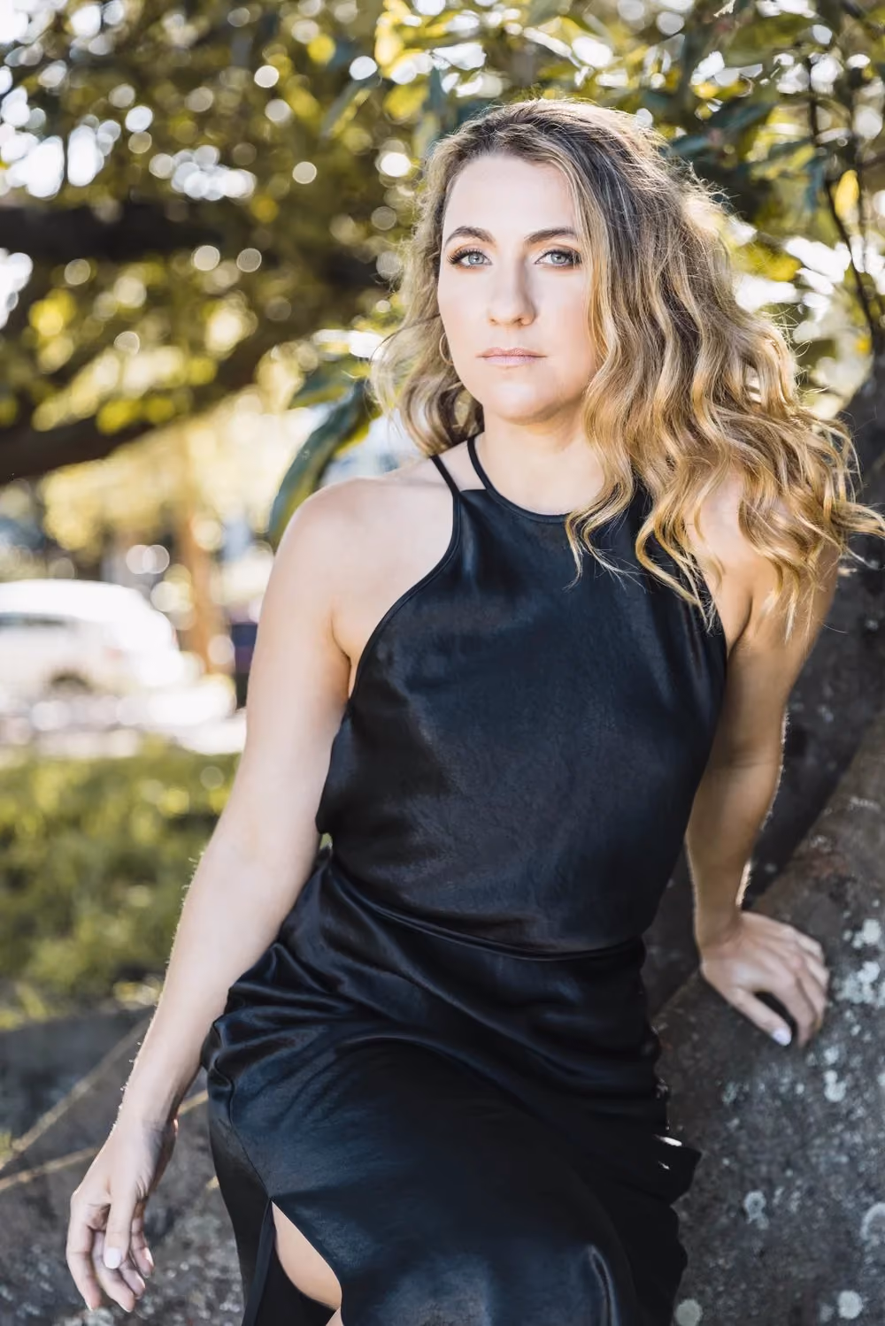 Blonde woman with wavy hair in a black dress sitting outdoors by a tree in dappled sunlight.