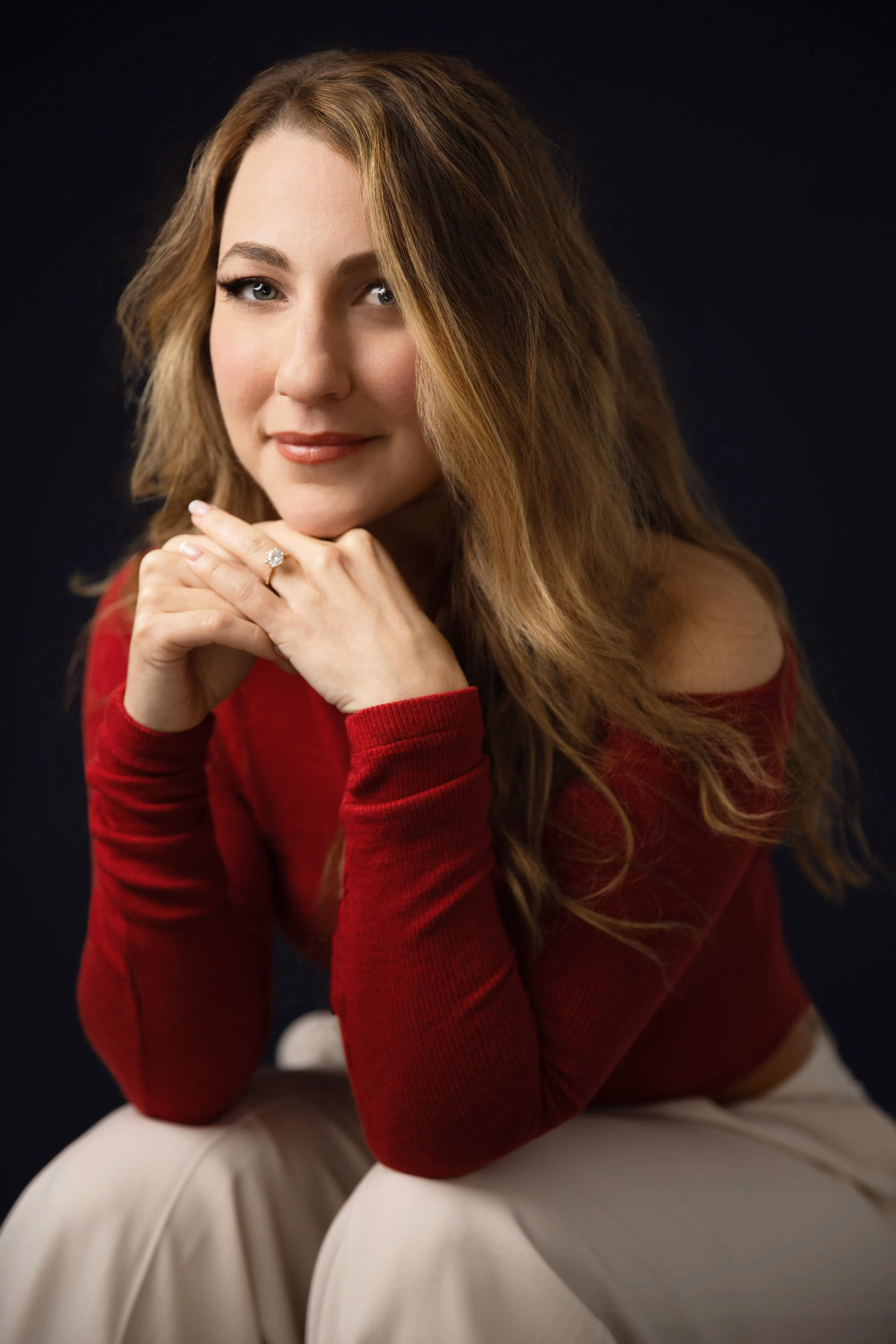 Woman with long blonde hair wearing red top and beige pants, resting her chin on clasped hands.