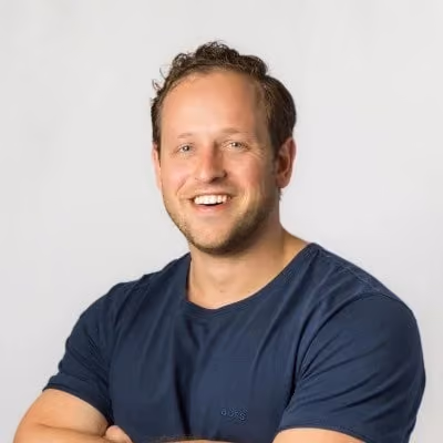 Smiling man with short curly hair wearing a navy blue t-shirt against a plain background.