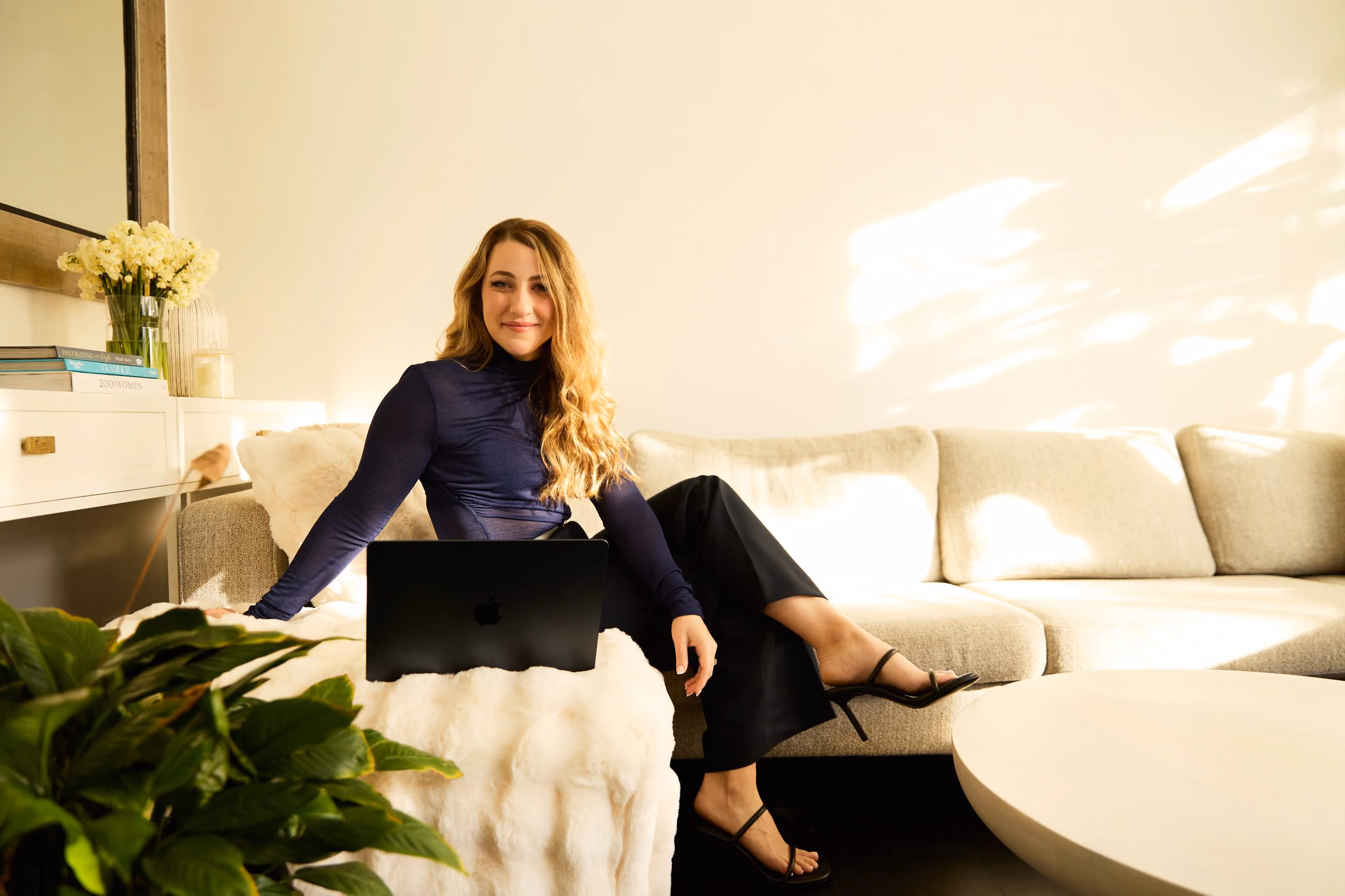 Smiling woman with long hair sitting on a beige couch with a laptop in a bright room.