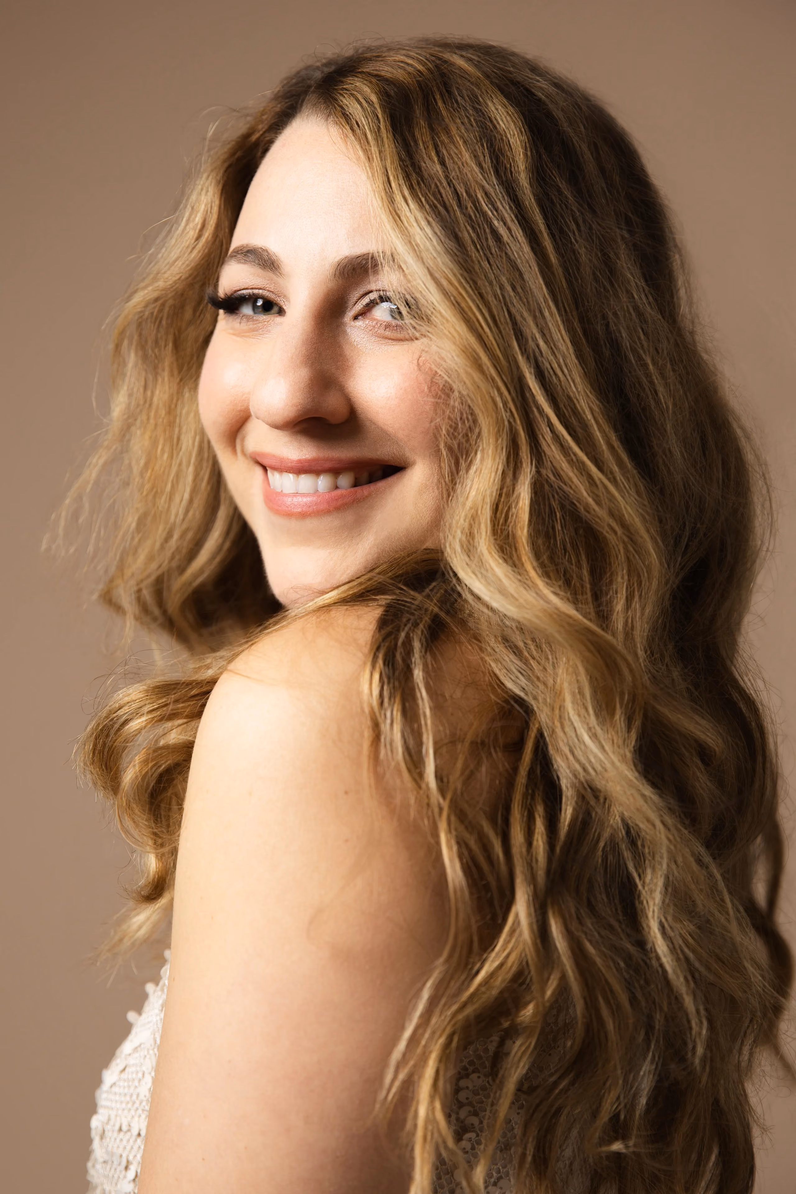 Smiling woman with long wavy blonde hair looking over her shoulder against a beige background.