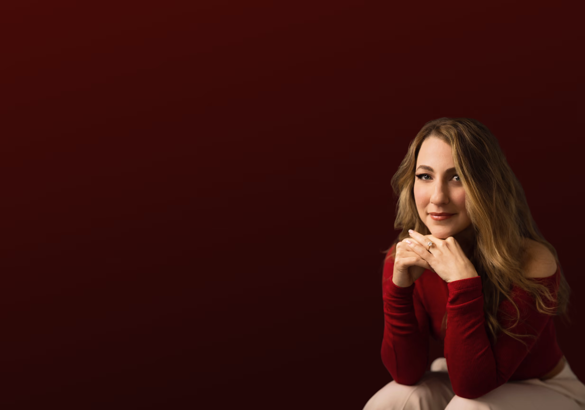 Smiling woman with long blonde hair in a red top resting her chin on her hands against a dark background.