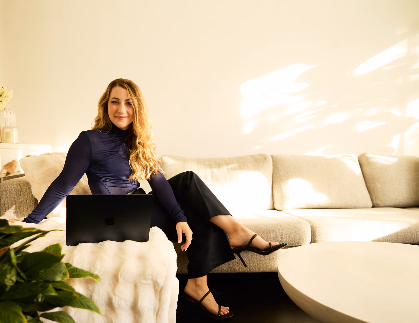 Woman smiling with long wavy hair sitting on couch using a black laptop in bright modern living room.