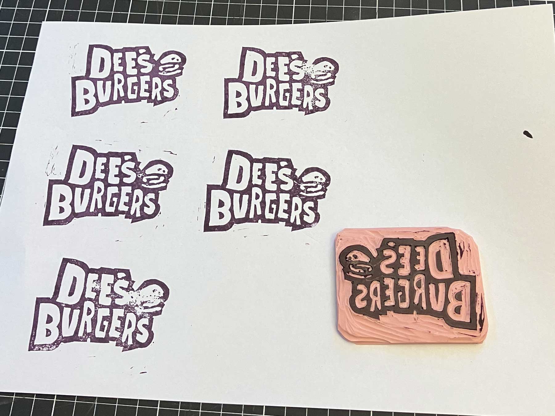 Five test stamps of the Dee's Burgers logo on white paper alongside the carved rubber stamp block, showing the refinement process
