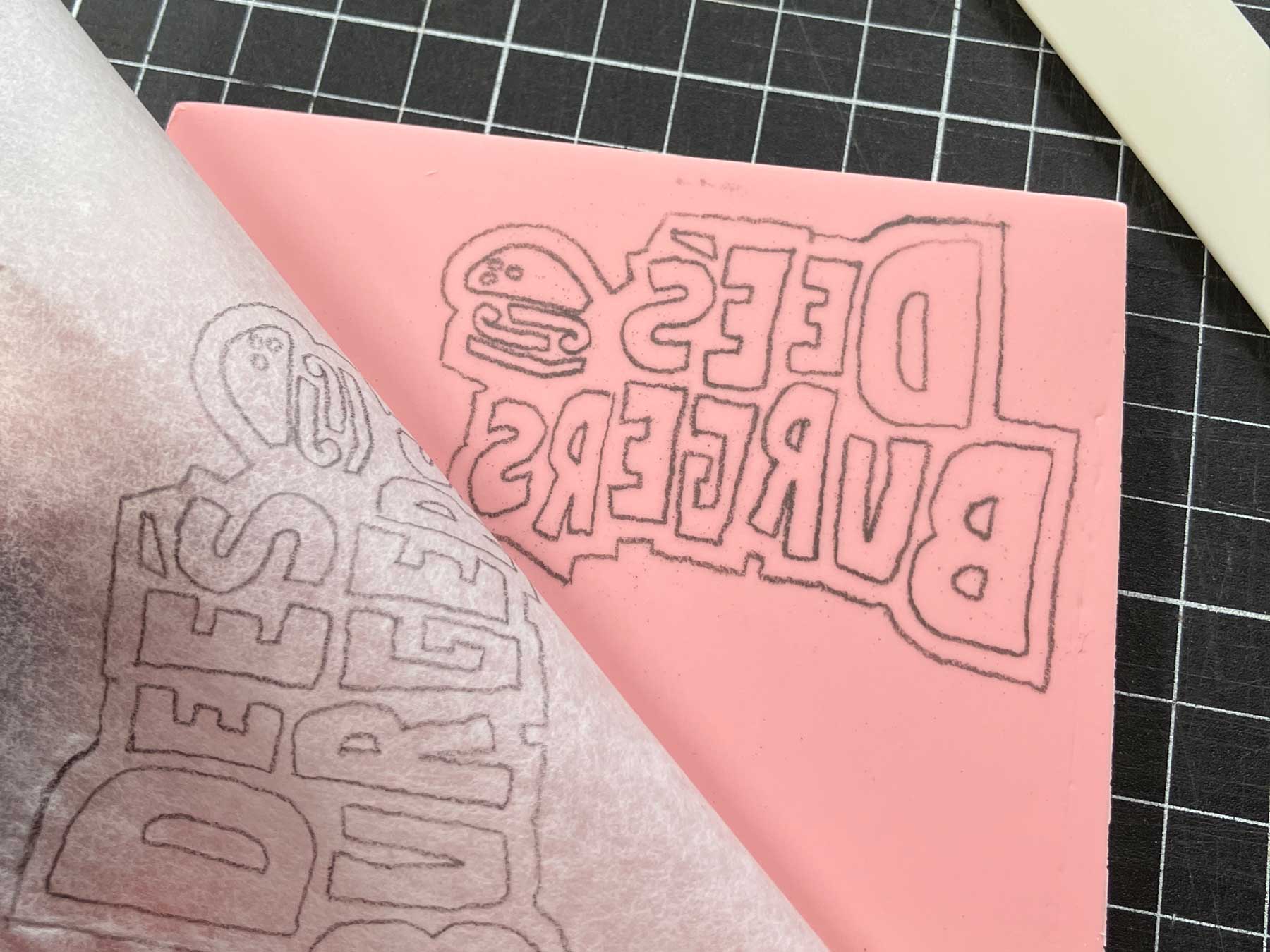Baking paper being peeled back from a pink rubber carving block to reveal the transferred Dee's Burgers logo outline in pencil