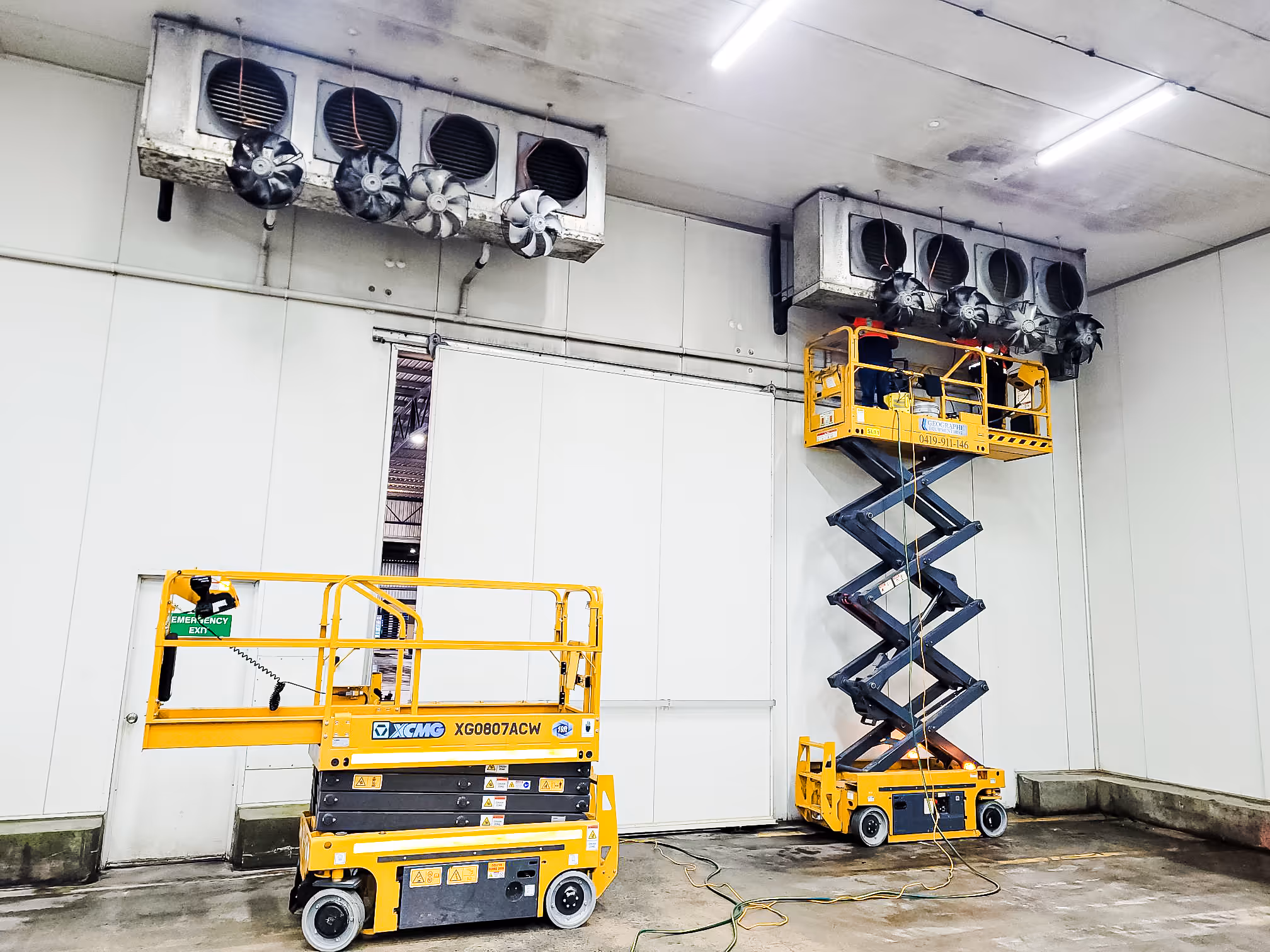 Two yellow scissor lifts inside an industrial space, one extended with workers servicing large ventilation fans mounted near the ceiling.
