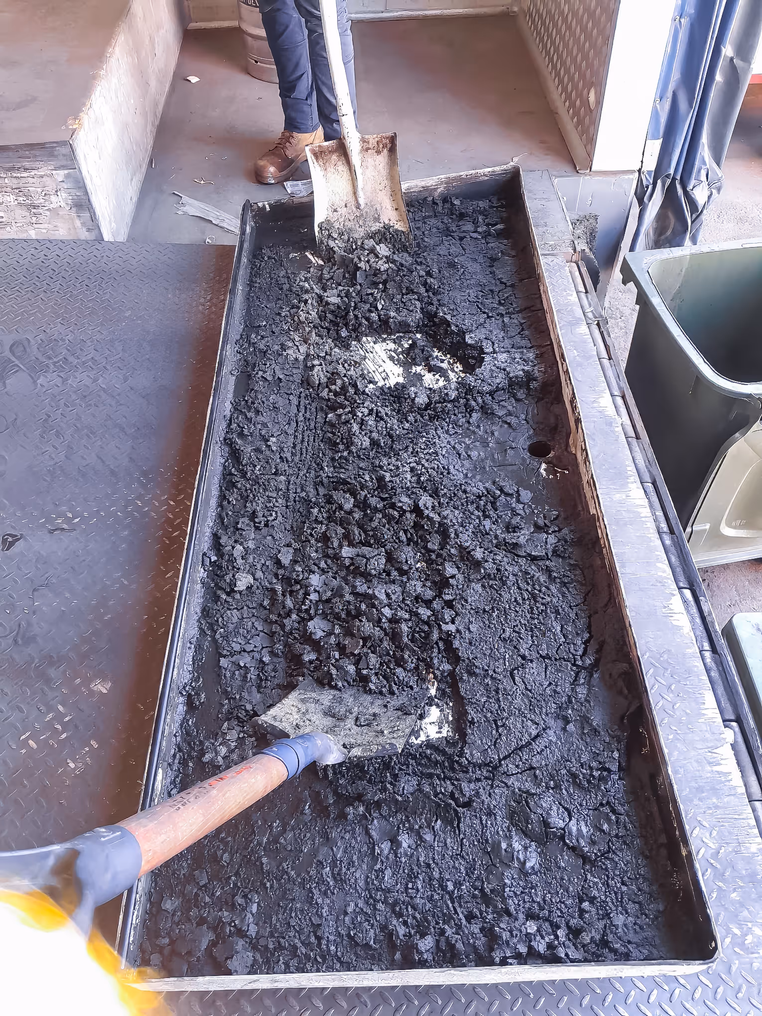 Two shovels mixing and moving wet black asphalt or tar in a large rectangular metal tray indoors.