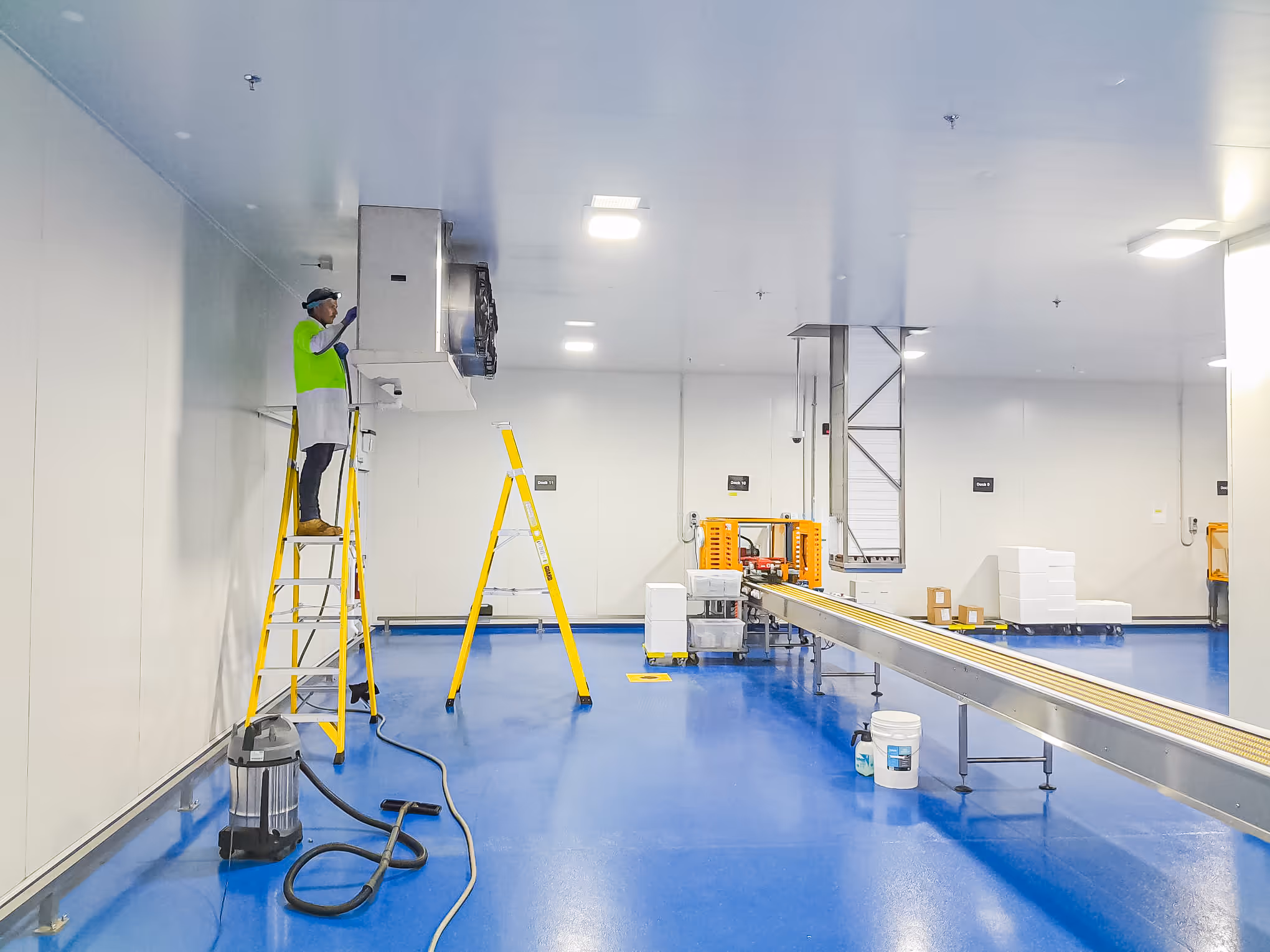 Worker in safety gear standing on a yellow ladder inspecting equipment on the wall inside a clean, blue-floored industrial facility.