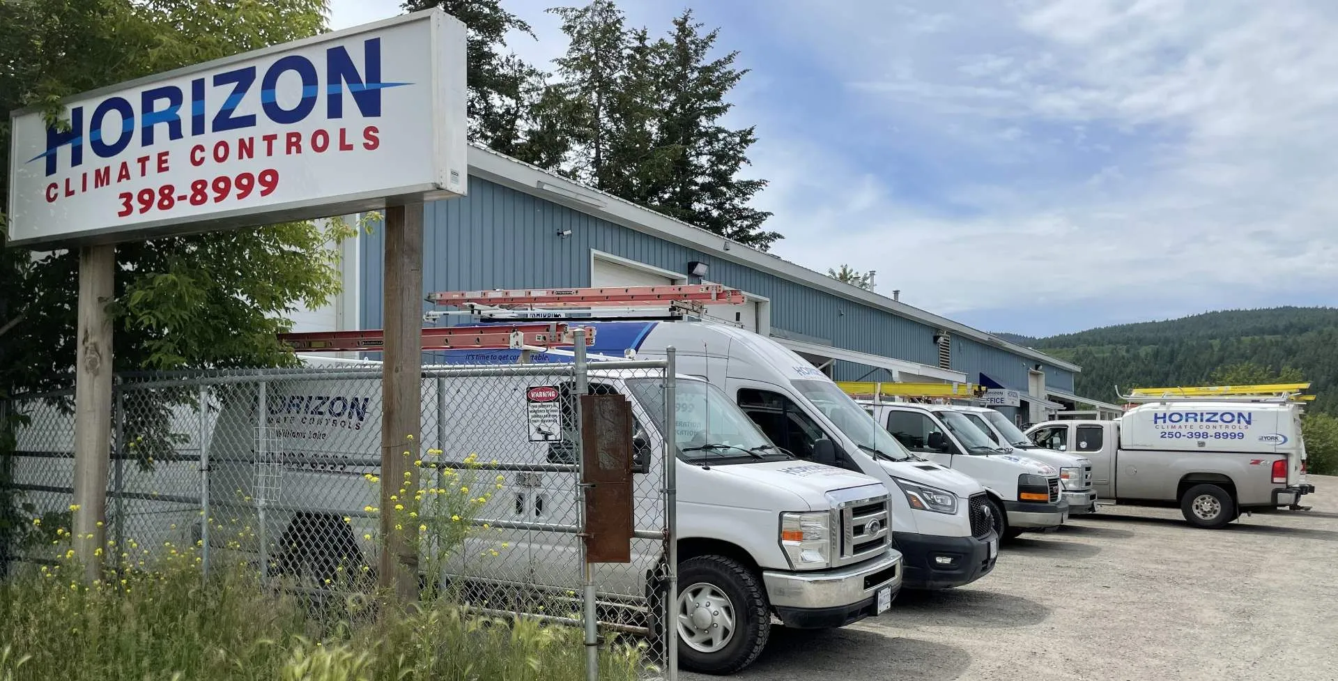 Row of white Horizon Climate Controls service vans parked outside a blue industrial building with trees behind.
