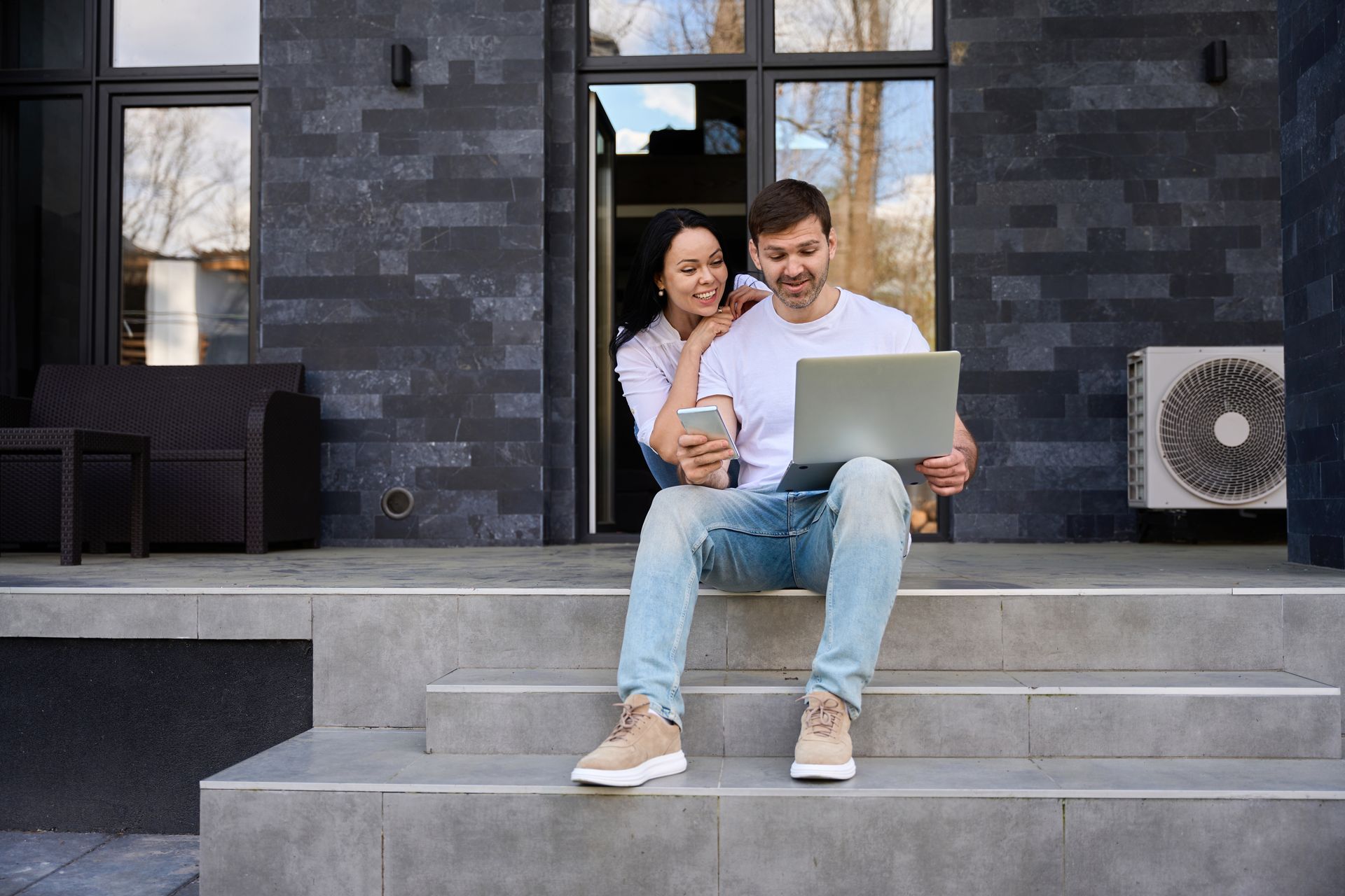 Couple sitting on steps outside a modern house, looking at a laptop and smartphone together.
