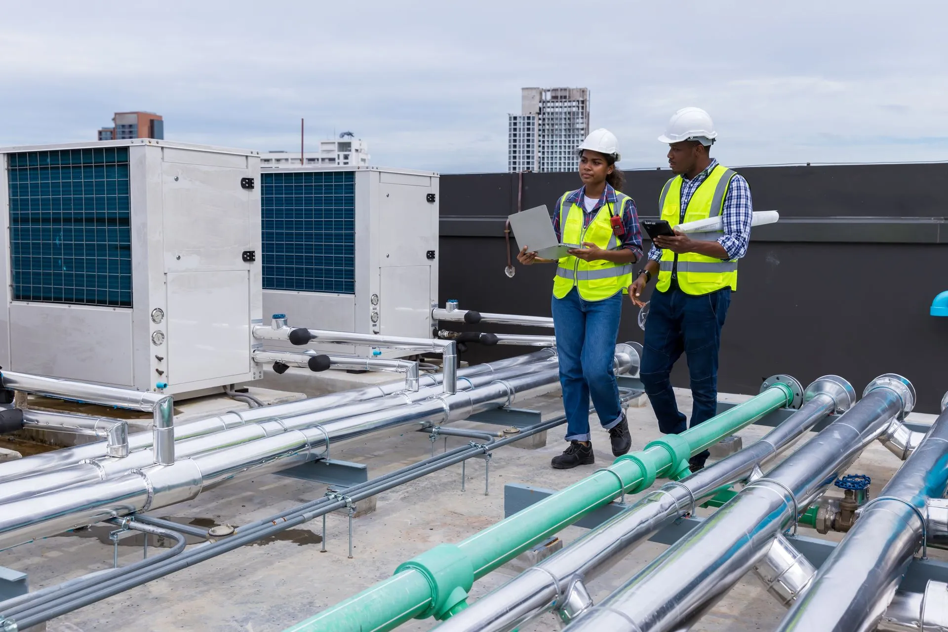 Two engineers in safety vests and helmets inspecting rooftop HVAC units and pipes with a laptop and tablet.