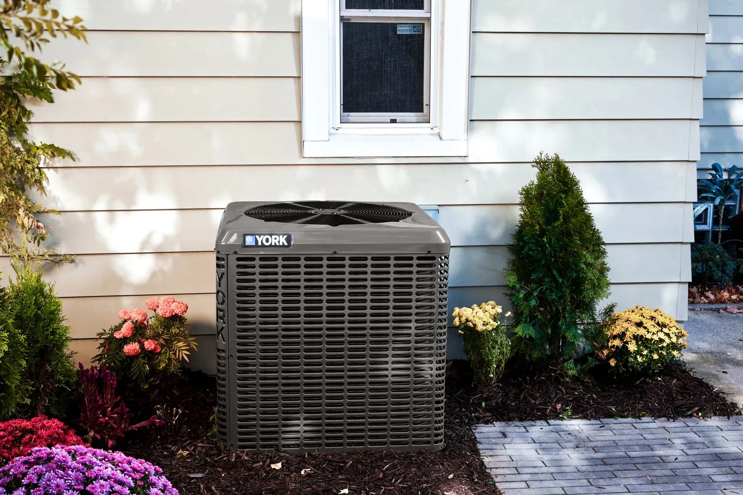 York air conditioning unit installed outside a beige house with surrounding flowers and shrubs.