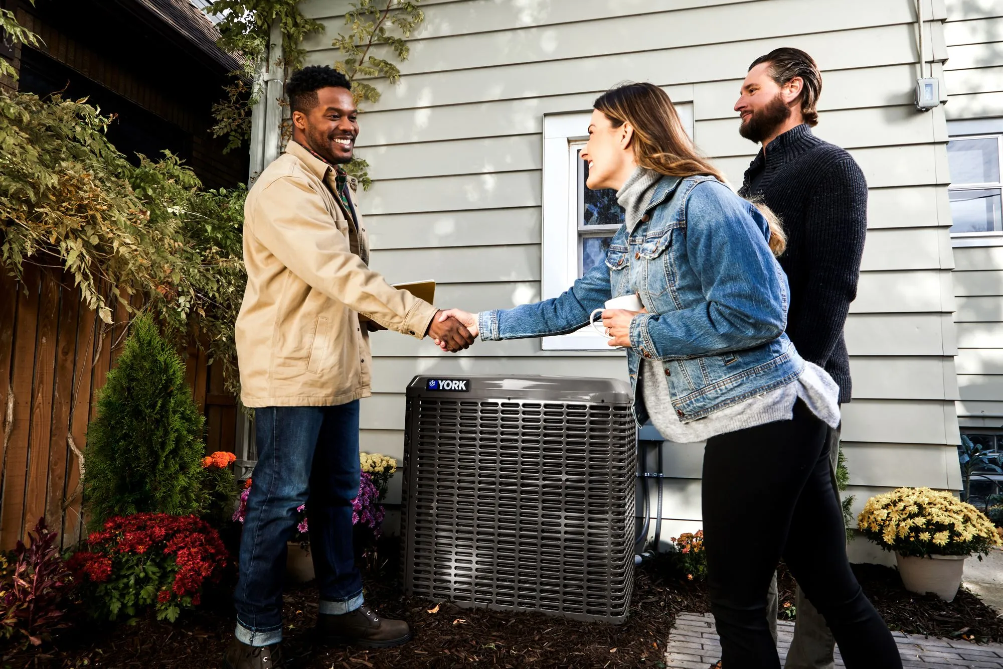 Woman shaking hands with a man near a York air conditioning unit outside a house, another man watches.