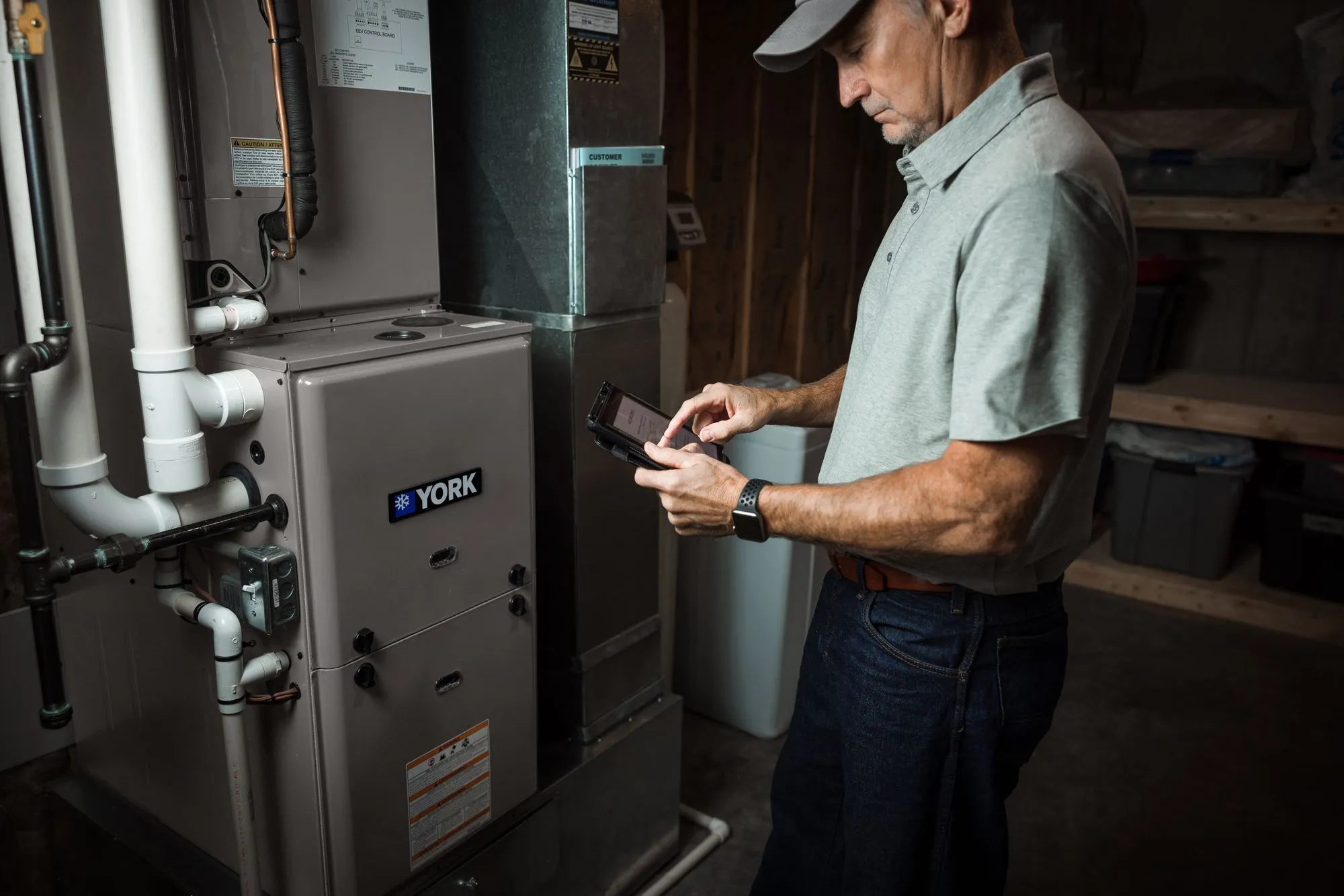 Technician in gray shirt and cap checking settings on a tablet near a York furnace unit.