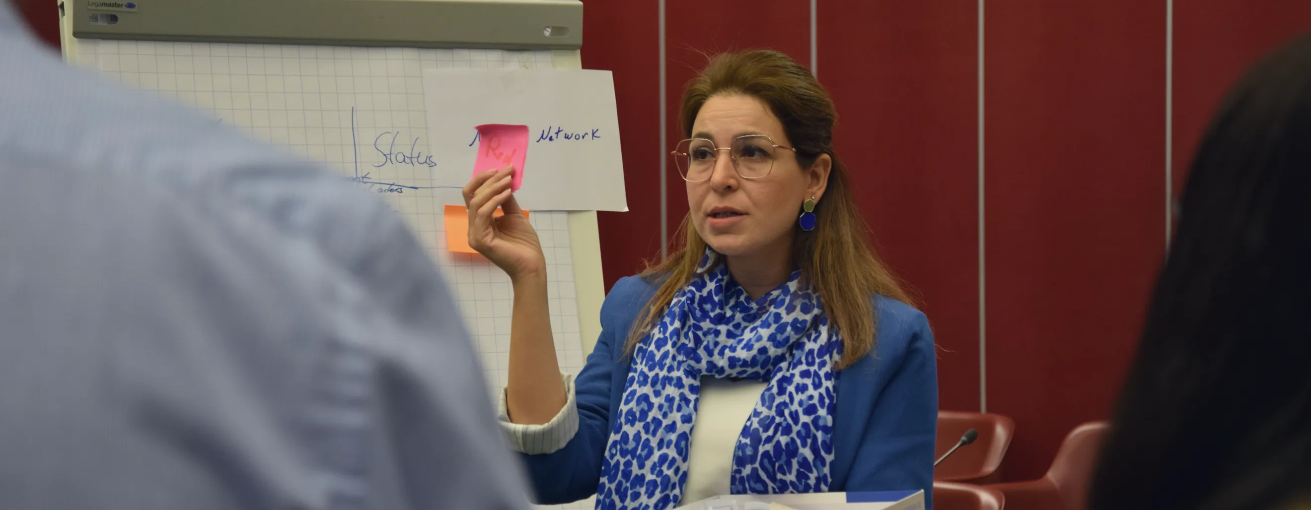 Two women in conversation, one holding a pink note, appearing to network or chat at a professional event or conference.