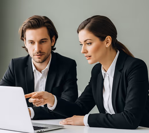 Two business professionals in suits looking at a laptop, with the woman pointing at the screen.