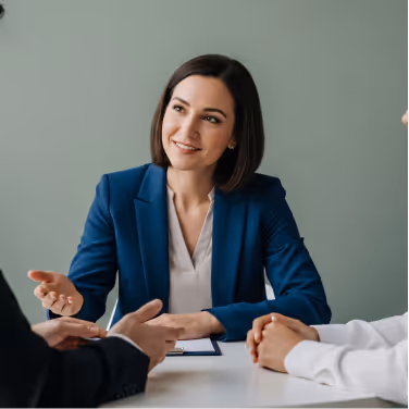 Smiling businesswoman in a blue blazer engaging in a discussion with two people across a table.