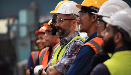 Group of diverse construction workers wearing hard hats and safety vests, standing in a row and looking forward attentively.