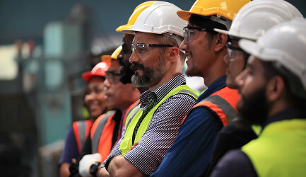 Group of diverse construction workers wearing hard hats and safety vests, standing in a row and looking forward attentively.