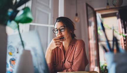 Woman wearing glasses and a mauve shirt sitting thoughtfully in front of a laptop in a softly lit room.