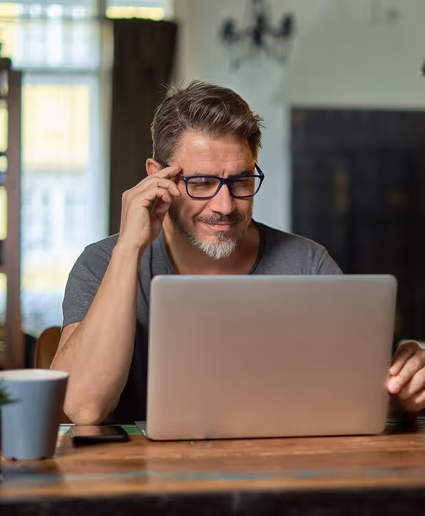 Middle-aged man wearing glasses, sitting at a wooden table and working on a laptop with a smartphone and a coffee cup nearby.