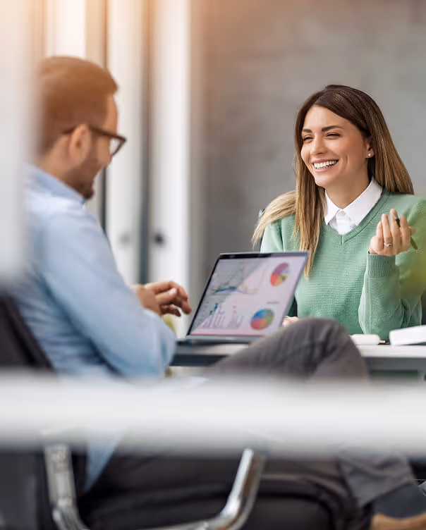 Two colleagues smiling and discussing data charts displayed on a laptop in a bright office.