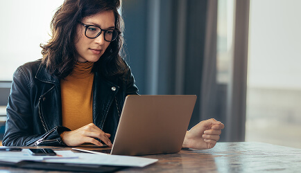 Woman wearing glasses and a leather jacket using a laptop at a table with papers and a phone.