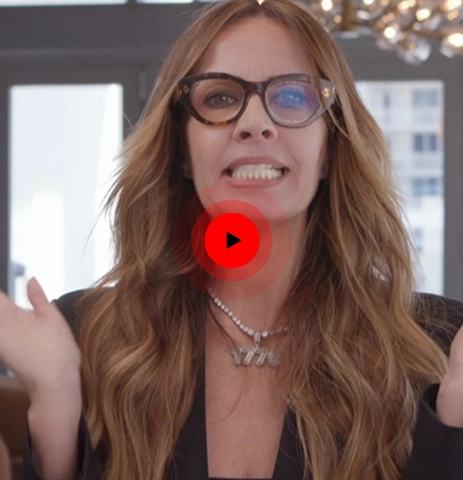 Woman with long wavy hair and glasses speaking indoors with hands raised near her head.